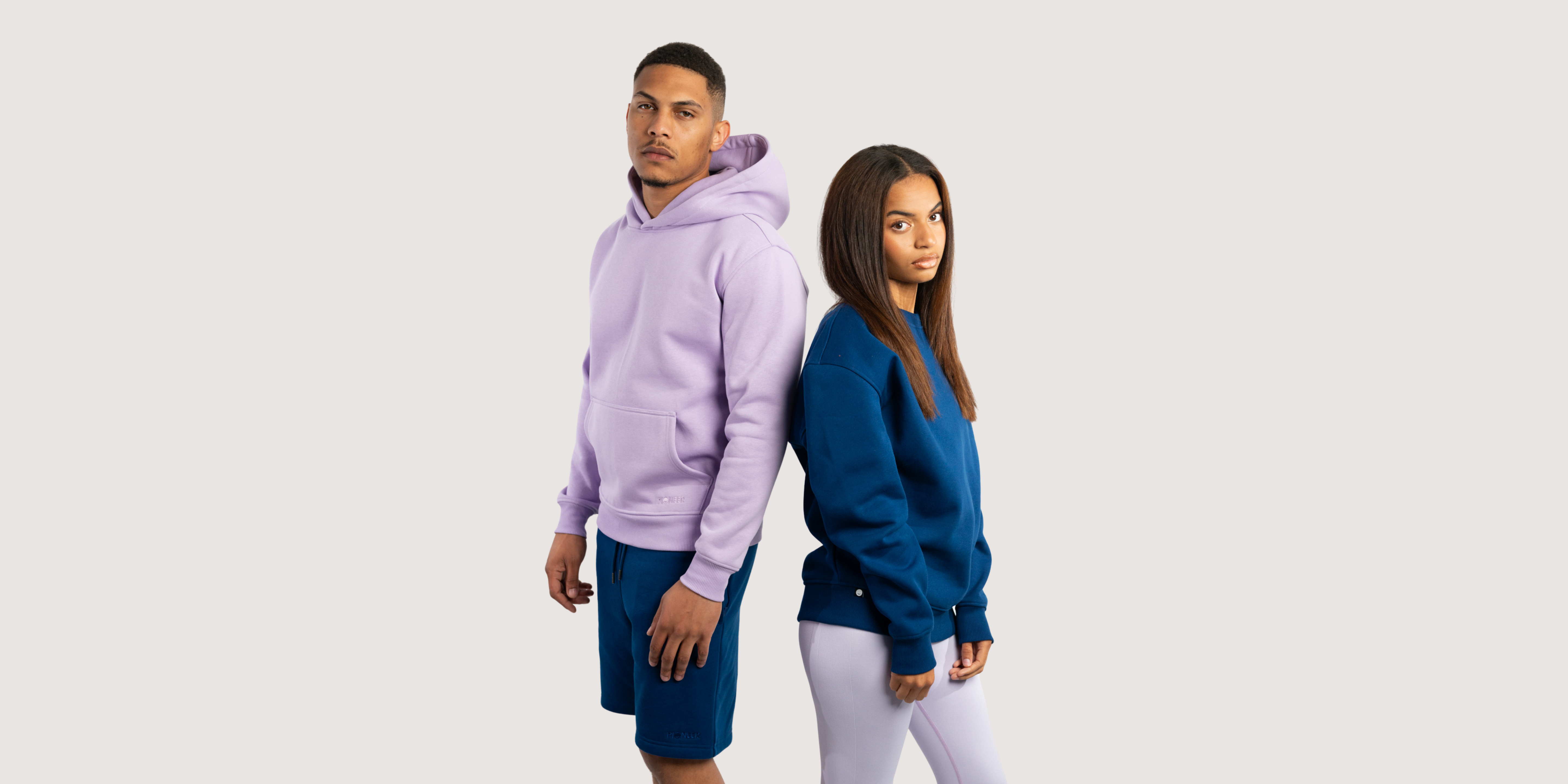 Model posing side to side. Male model wearing Orchid Petal hoodie with  Navy Shorts and Female model wearing Navy Crew Neck Sweatshirt and Orchid Petal Leggings. All the products are made from sustainable eco-friendly materials.