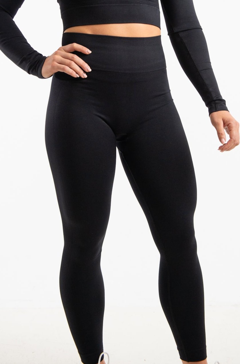 This is a full length image which shows a model wearing a matching set which include a cropped fitted long sleeve top in the shade Black , she is wearing this with a pair of high waisted seamless leggings in the same shade which are made from sustainable eco-friendly materials. The clothes are form fitting which helps to show the models athletic build. The model is standing against a plain white background with her right hand on her hip.