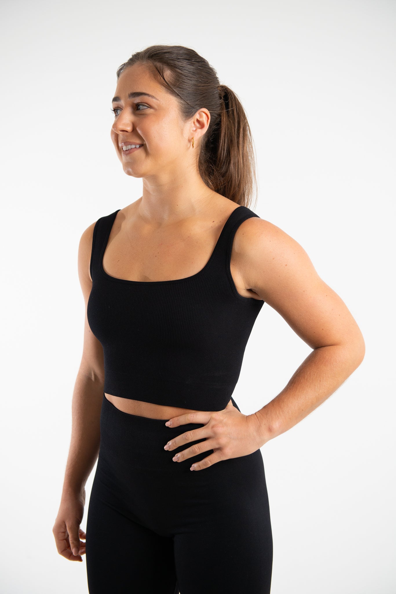 This image shows a woman wearing our seamless fitted vest top in Black which is a cropped top with a square neckline, she is also wearing a matching pair of high waisted shorts also in Black which are made from sustainable eco-friendly materials. She is posing with one hand on her hip against a plain white background.