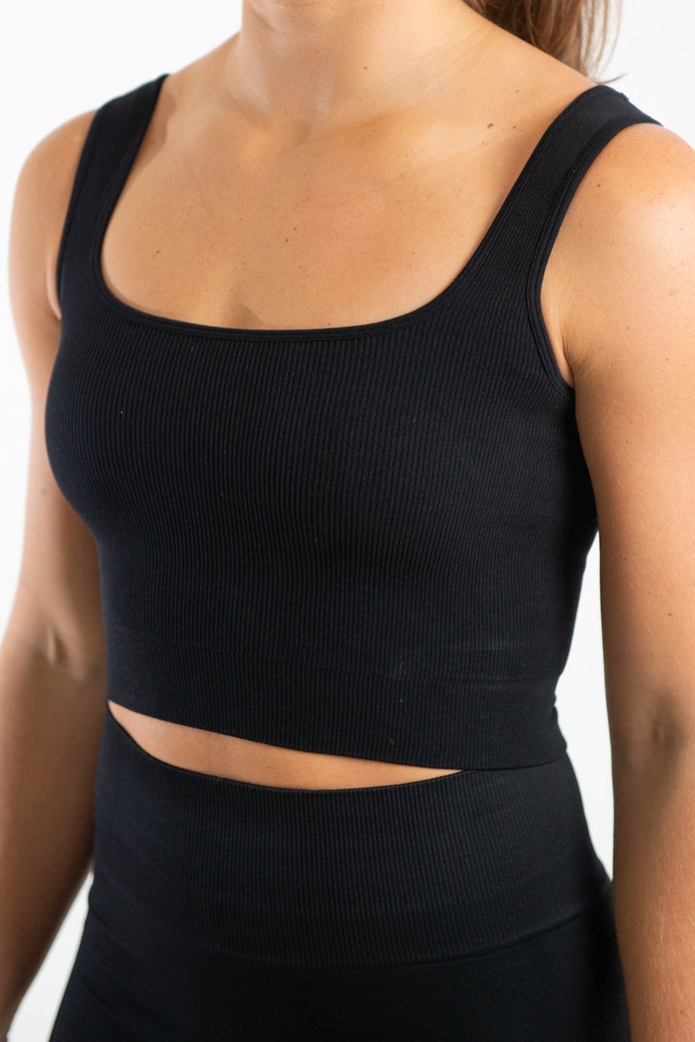 This image shows a woman wearing our seamless fitted vest top in Black which is a cropped top with a square neckline, she is also wearing a matching pair of high waisted shorts also in Black which are made from sustainable eco-friendly materials.