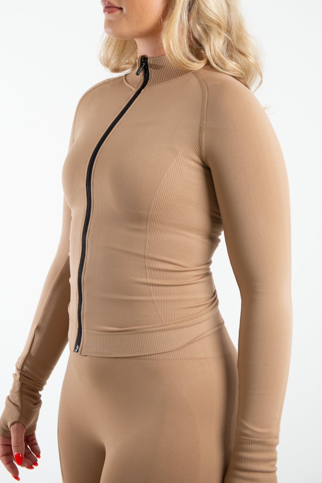 A woman standing to the side against a neutral white background with her arms positioned to the side of her. She is wearing a long sleeve zip fitted top in the shade Hazelnut it has a high neckline this is paired with a matching pair of leggings which are both made from sustainable eco-friendly materials.