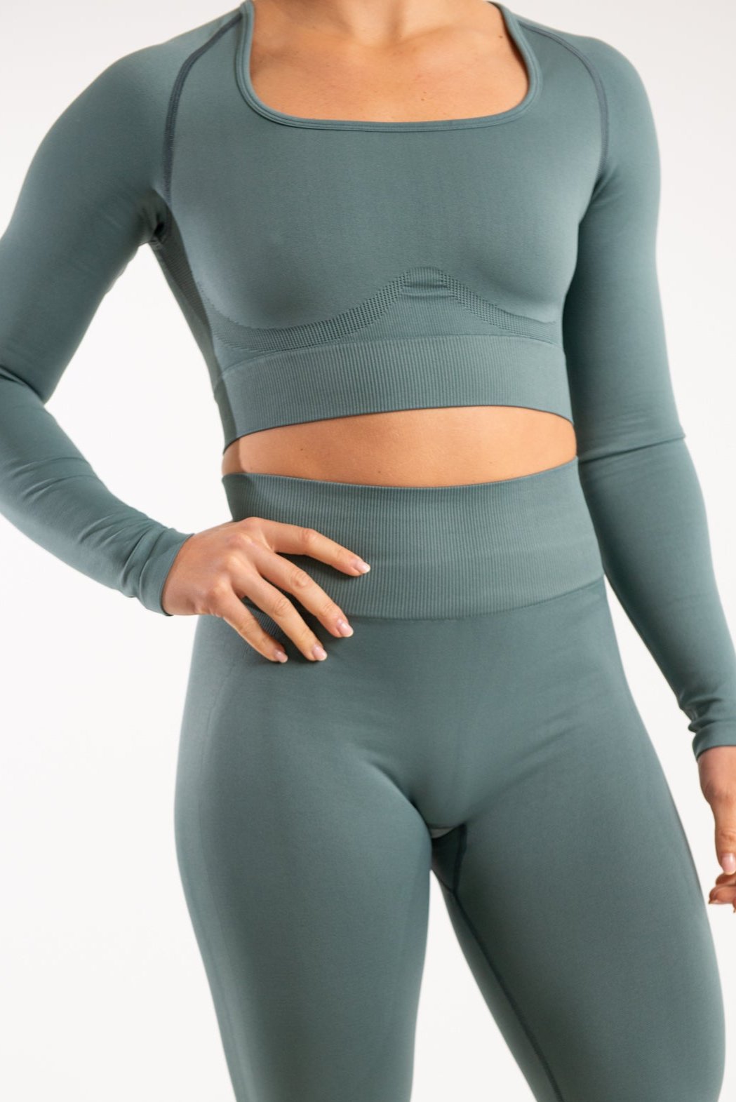 A woman facing forward wearing a long sleeve crop top with a high square neckline paired with matching high waisted leggings which are both made from sustainable eco-friendly materials and are in the shade Dark Forest Green. The woman has an athletic build and is standing against a plain white background with her arms to her side.