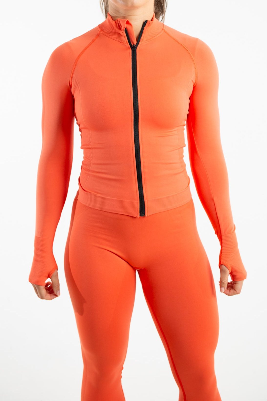 A woman standing with her arms swaying to the side of her. The woman is wearing a seamless form fitted top with a zip running through the middle in the shade Mandarin Red. She is also wearing a matching pair of leggings which are both made from sustainable eco-friendly materials. The woman is standing against a white background.