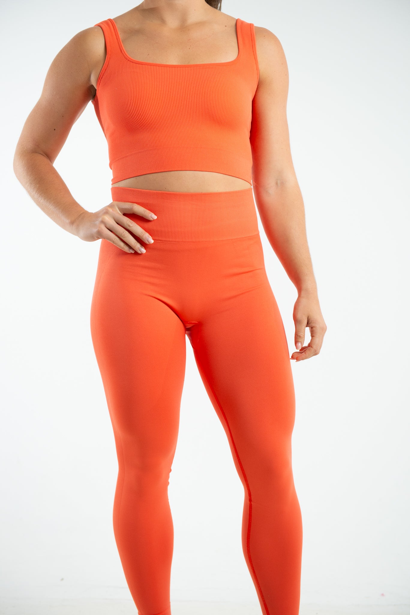 This is a full length image which shows a model wearing a matching set which includes a cropped fitted vest top in the shade Mandarin Red which has a square neckline, she is wearing this with a pair of high waisted seamless leggings in the same shade which are made from sustainable eco-friendly materials. The clothes are form fitting which helps to show the models athletic build. The model is standing against a plain white background with her hight hand on her hip.