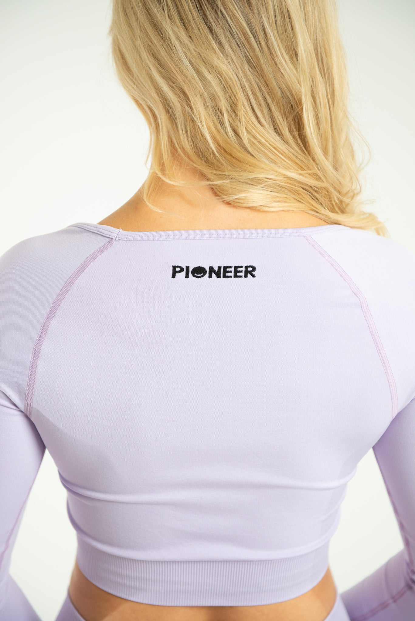 A close up back image of a woman wearing a long sleeve seamless fitted crop top. At the top of the crop top it contains the Pioneer logo in a black font. The top is made from sustainable eco-friendly materials, and is in the shade Orchid Petal. The model is stood Infront of a plain white background.
