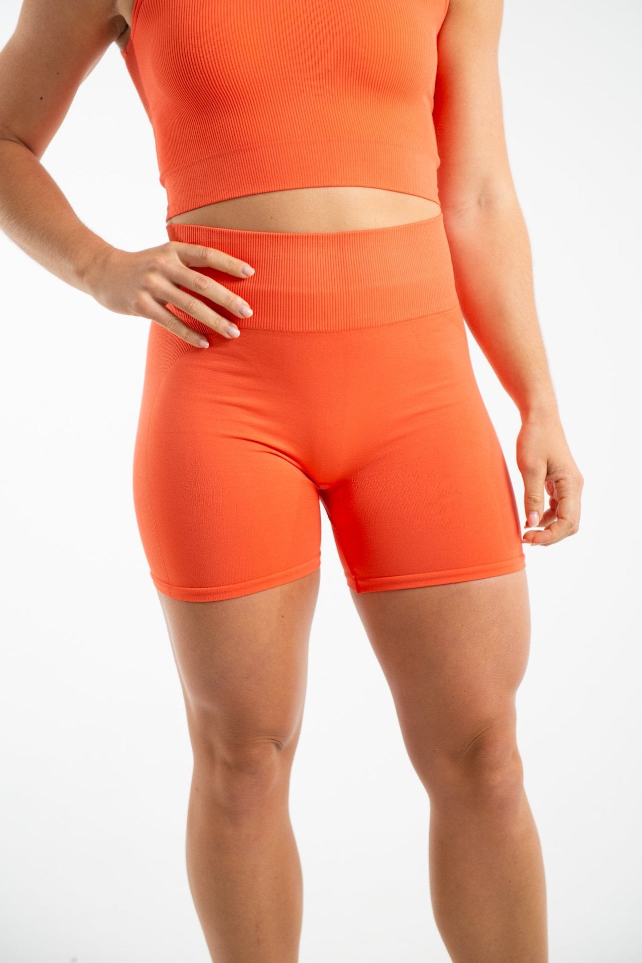 A shot of a woman wearing a pair of seamless cycling shorts in the colour Mandarin Red. The shorts have a high waist band and are made from a stretchy eco-friendly material and come down to the middle of her thigh. The woman has a very athletic build and is standing against a white background with her right hand placed on her hip.